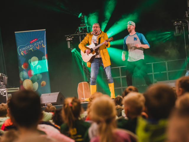 Bremi steht in einem gelben Regenmantel, Gummistiefeln und Gitarre auf der Bühne und singt den Song "Pfütze hüpfen"